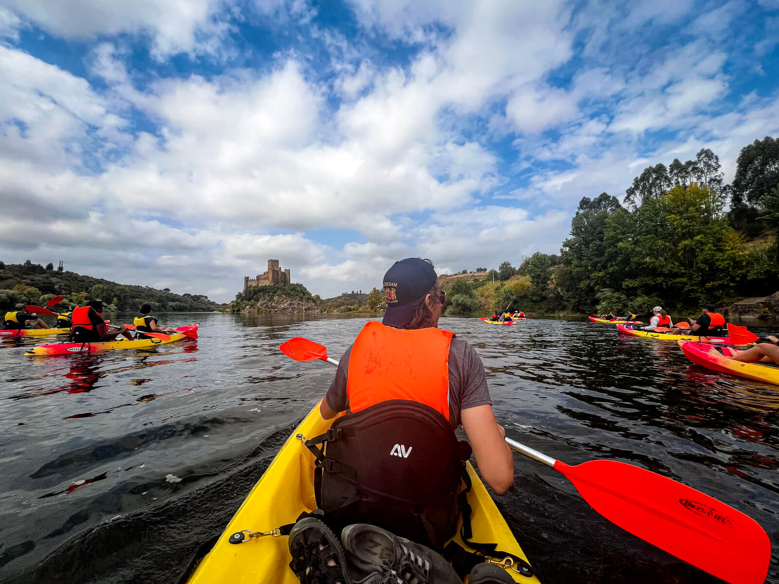 A person in an orange life vest and backwards baseball hat paddles a yellow kayak through a calm body of water, toward a castle perched on the distant shore. Other kayakers are visible on either side.