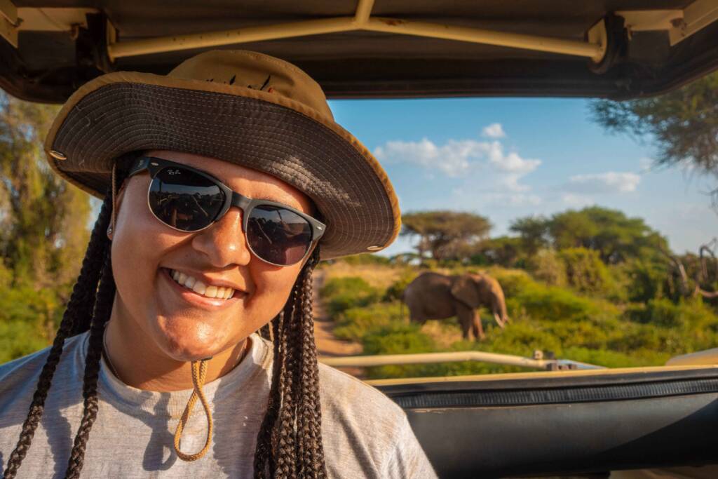 A person with long braids and a khaki safari hat sits in an open-sided vehicle, smiling at the camera. An elephant is visible in the background, surrounded by trees and scrub brush.