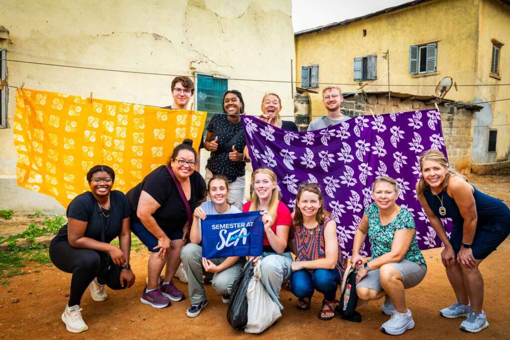 A group of about a dozen people poses in front of a clothesline featuring colorful batik-dyed cloths. Two people hold a small blue flag that says “Semester at Sea.”