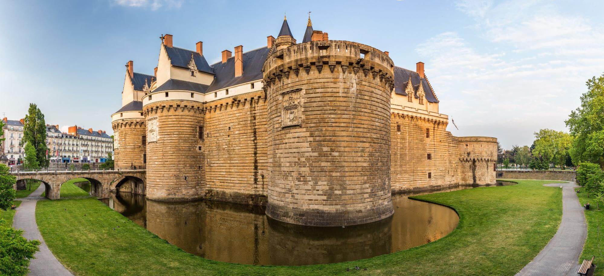 A panoramic view of a stone castle, the Château des Ducs de Bretagne, featuring medieval architecture with cylindrical stone towers, a surrounding moat, and a stone bridge.