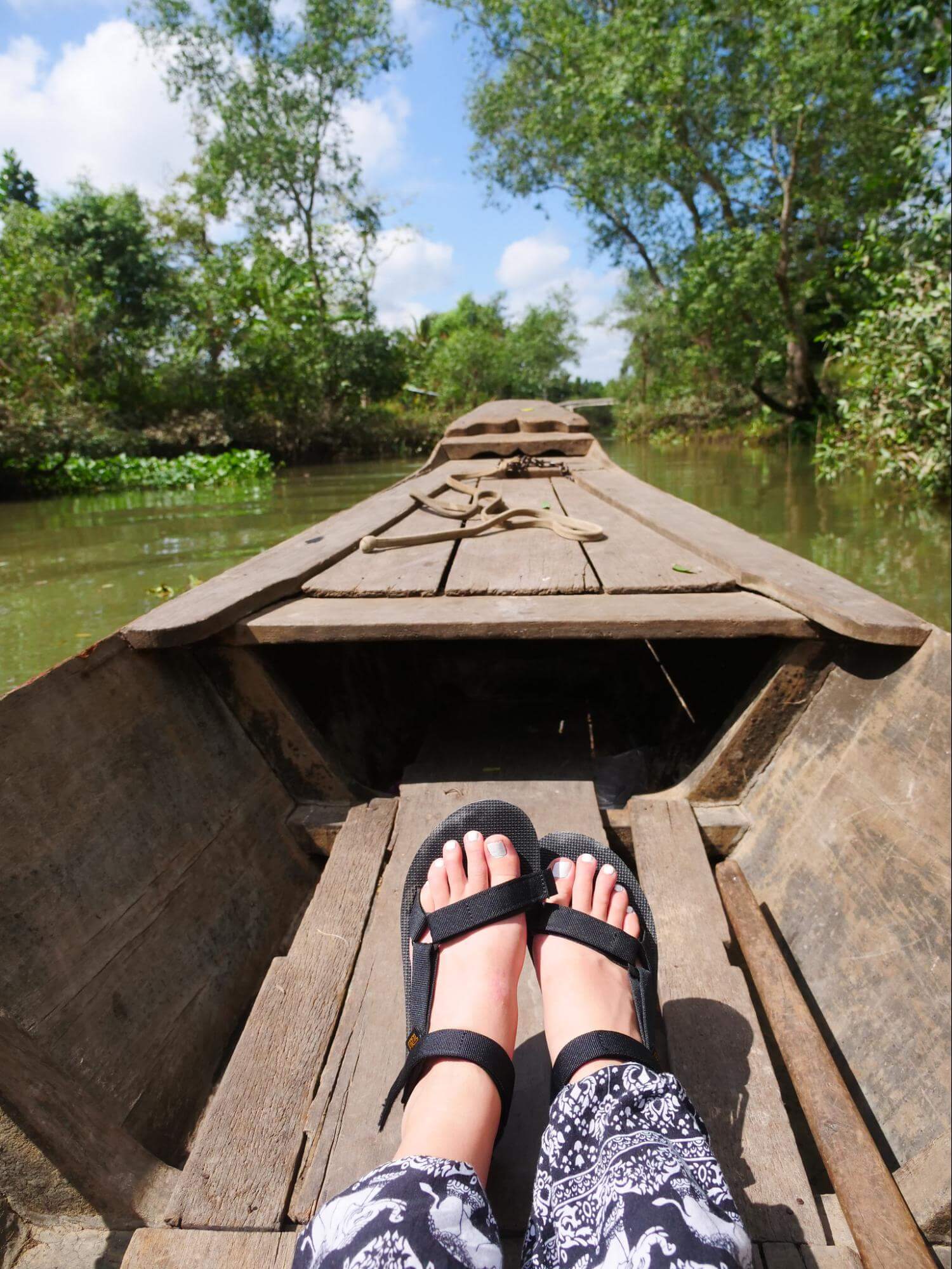 POV shot of a pair of sandaled feet, sitting in a wooden boat as it drifts in a calm, tree-lined body of water.