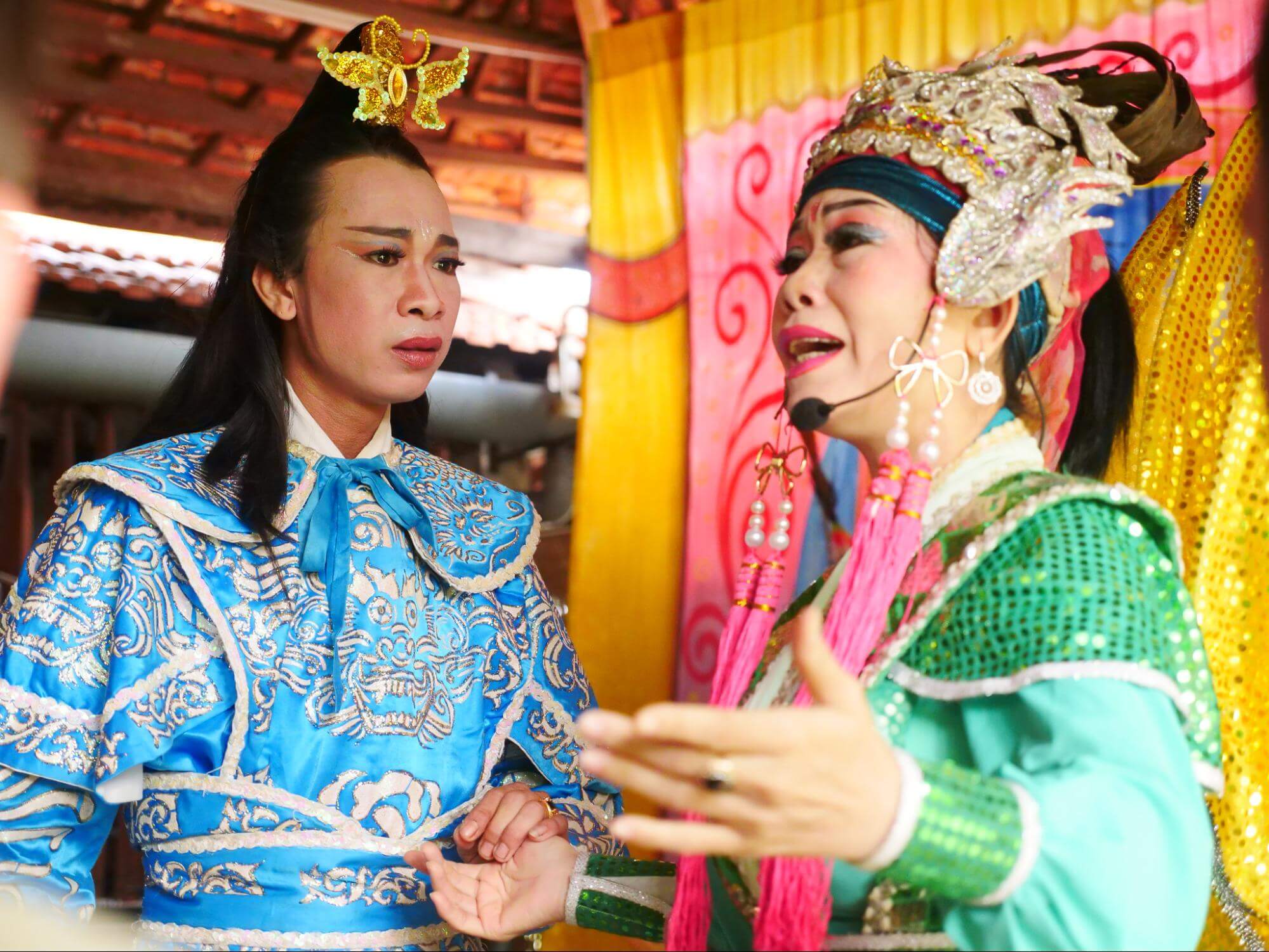 Two performers in colorful costumes and makeup interact onstage; one sings while the other listens.