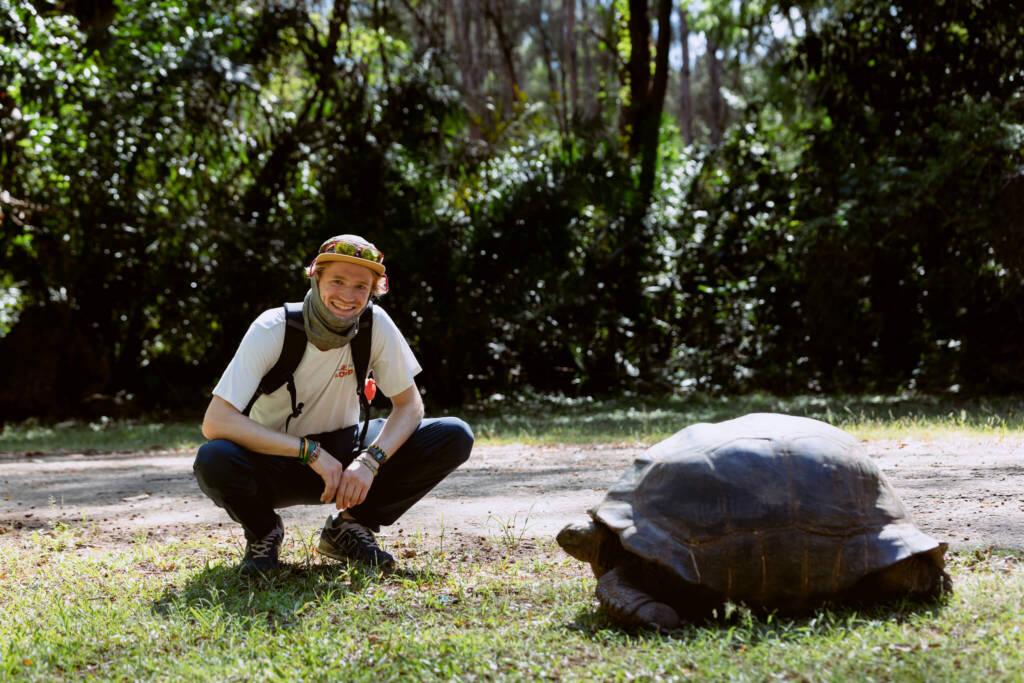 A young man crouches next to a giant tortoise.