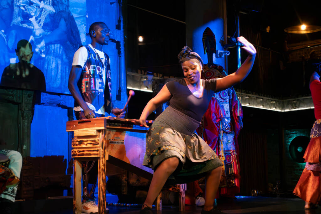 A performer in a flowered skirt and black headscarf dances onstage in front of musicians playing traditional South African instruments. 