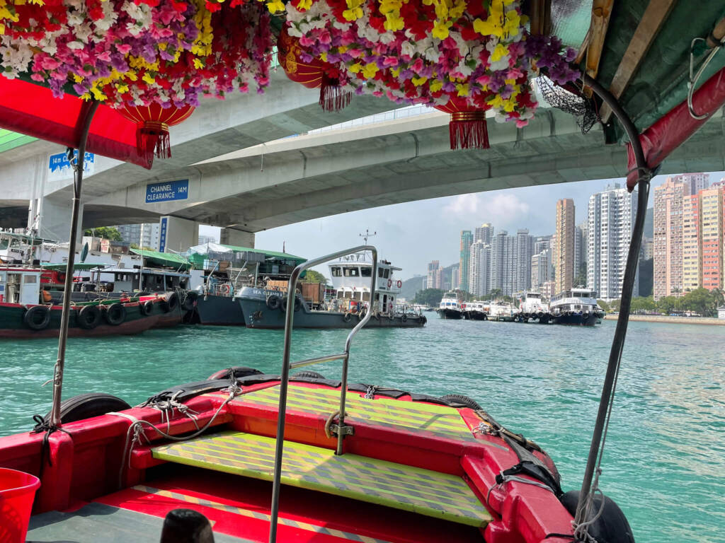 A view of skyscrapers is visible from underneath the canopy of a boat, lined with flowers and red silk lanterns. The boat sails through a busy harbor filled with other watercraft. 