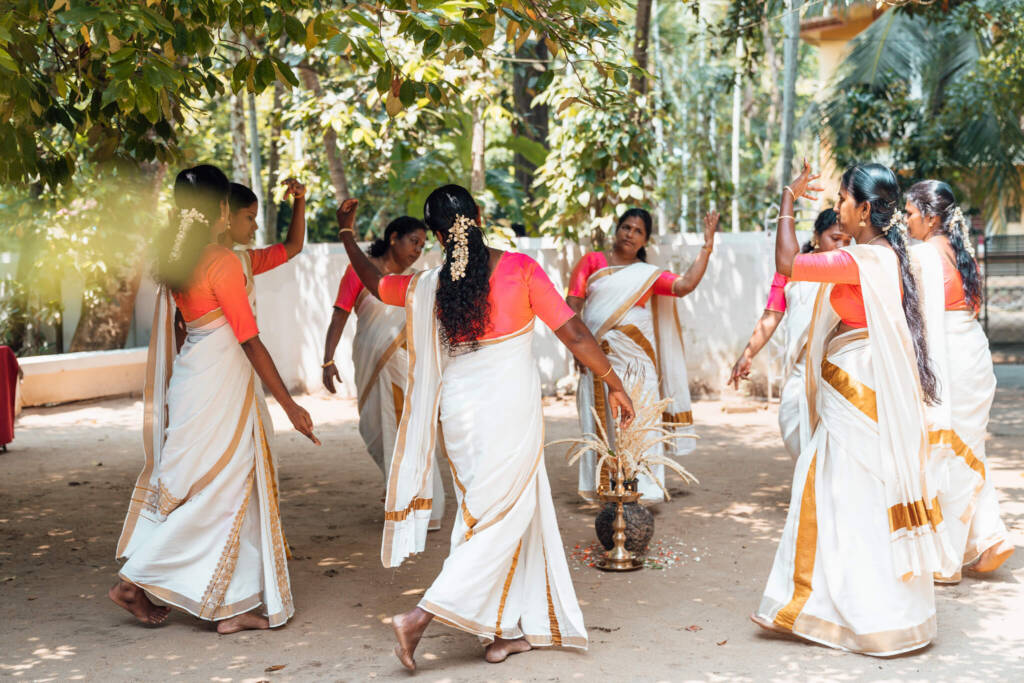 A group of women in pink, white, and gold saris dances in a circle around a golden candlestick and an urn filled with dried plant stalks. 