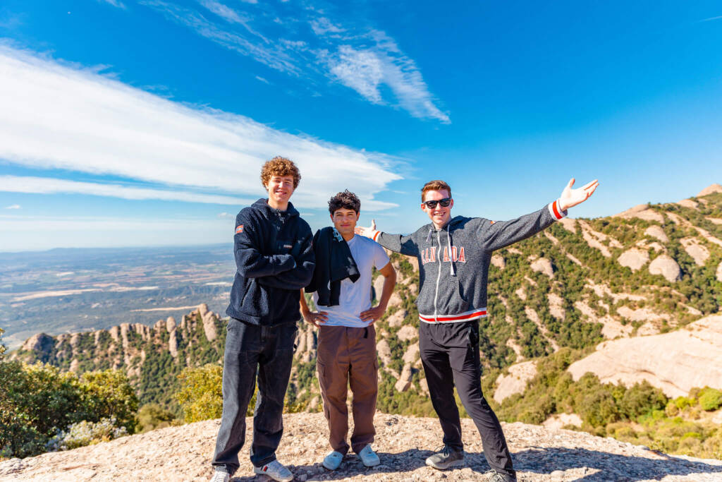 Three young men pose against a sprawling, sunny landscape of rocky outcroppings and green scrub brush. 