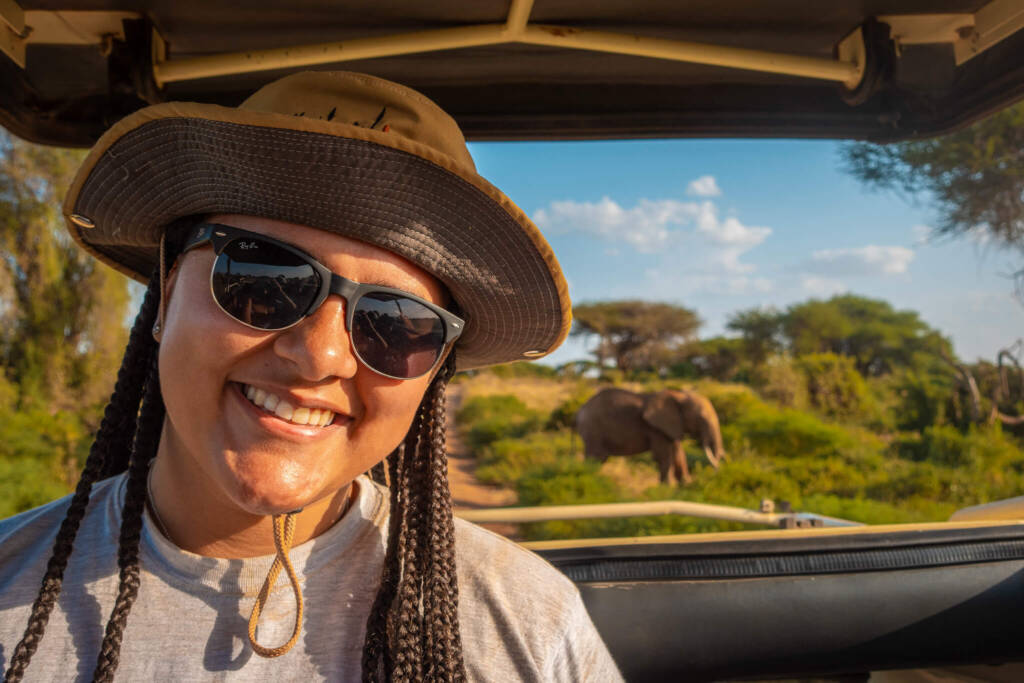 A student in a safari hat and sunglasses smiles at the camera while sitting in an open-air safari vehicle. An elephant is visible in the savannah behind them.