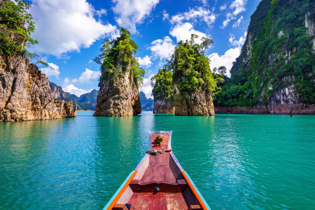 The view from the bow of a wooden boat floating on turquoise waters, heading toward towering limestone karsts covered with lush green vegetation. Rugged cliffs rise on either side against a vibrant blue sky with scattered white clouds.