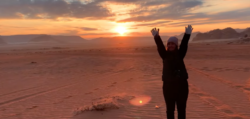A student stands in a desert against the sunset, waving their arms and smiling at the camera. 