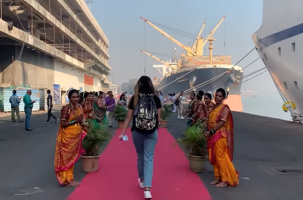 A student walks down a pink painted pathway in a ship’s port, with large ships visible next to the dock. Two rows of women in colorful saris stand on either side of the pathway.