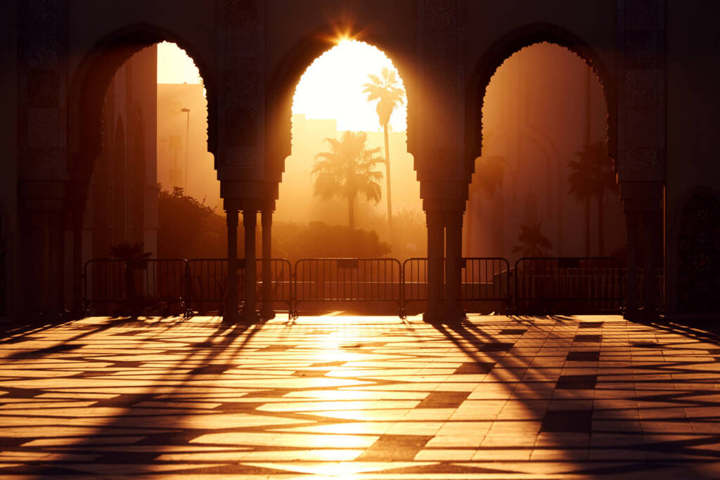 Hazy, late-afternoon sunlight streams through the ornate archways of a mosque in Casablanca. Palm trees are silhouetted against the sun. 