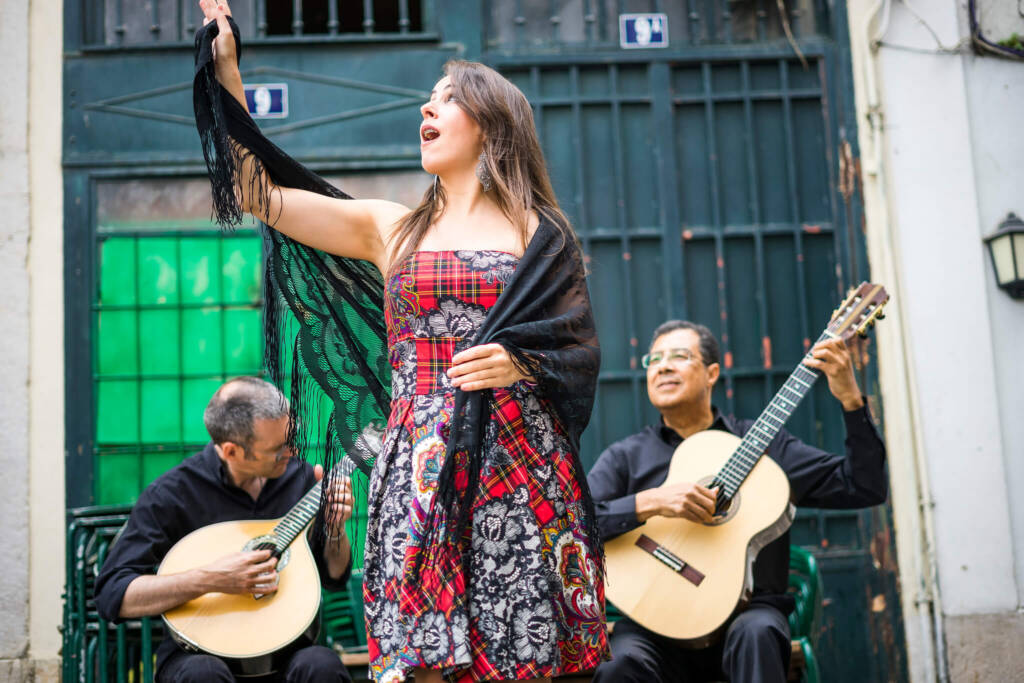 A woman in a red plaid dress and black lace shawl sings in front of two men playing string instruments on the street. 